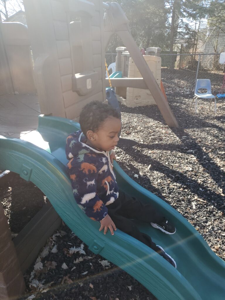 A young child sits on a slide at a playground, wearing a colorful animal-patterned sweater, ready to slide down into a gravel base.