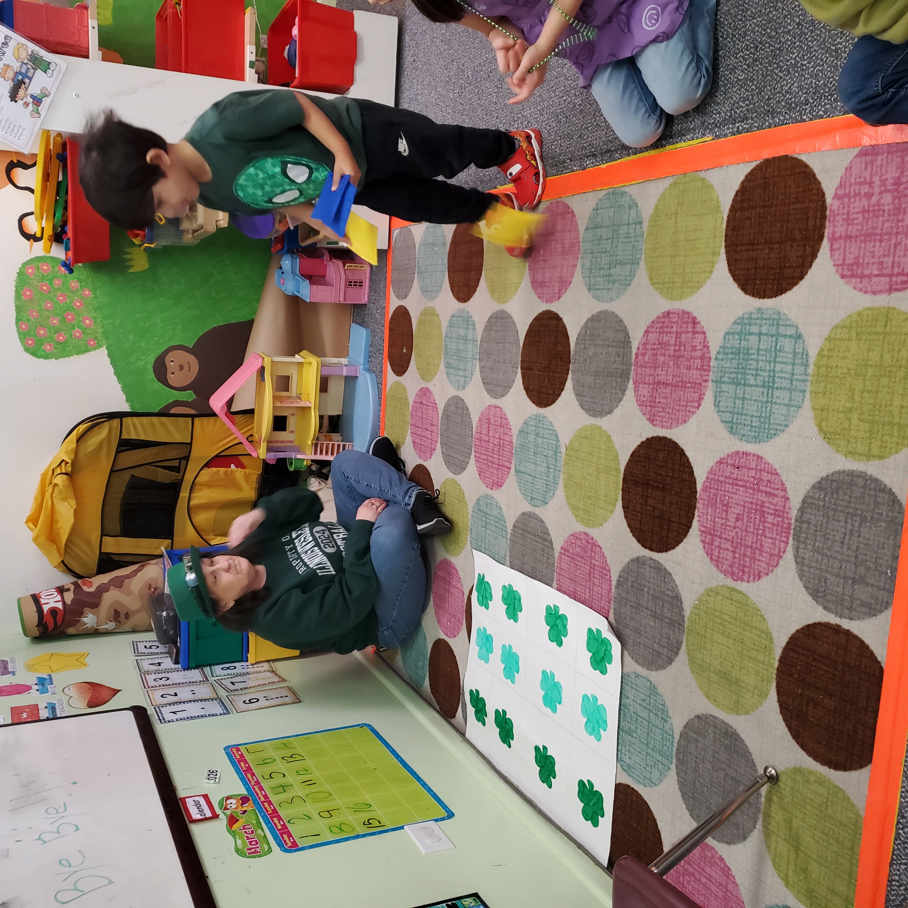 A teacher sits on a colorful carpet in a classroom, engaging with children as one boy plays with blocks, while others observe. Green shamrocks are visible on the floor, indicating a themed activity.