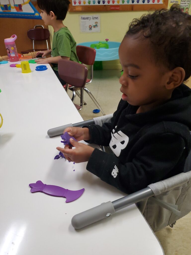 A young child focused on molding purple playdough shapes at a table, while another child plays with toys in the background.